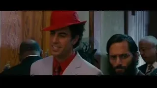man in red hat white suit walking hallway — The Dictator